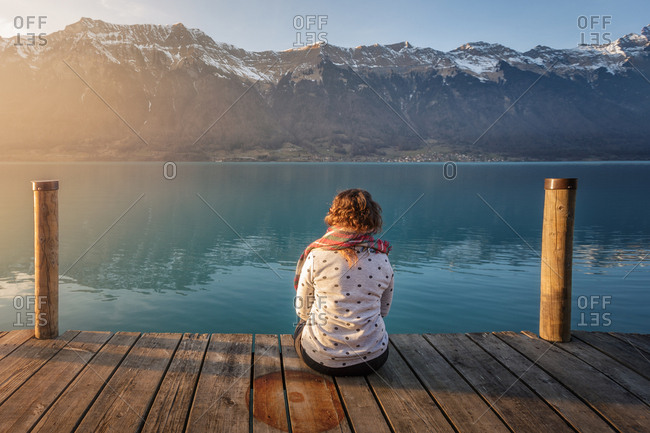 Back view of woman sitting on wood pier above turquoise lake in snowy mountains of Switzerland