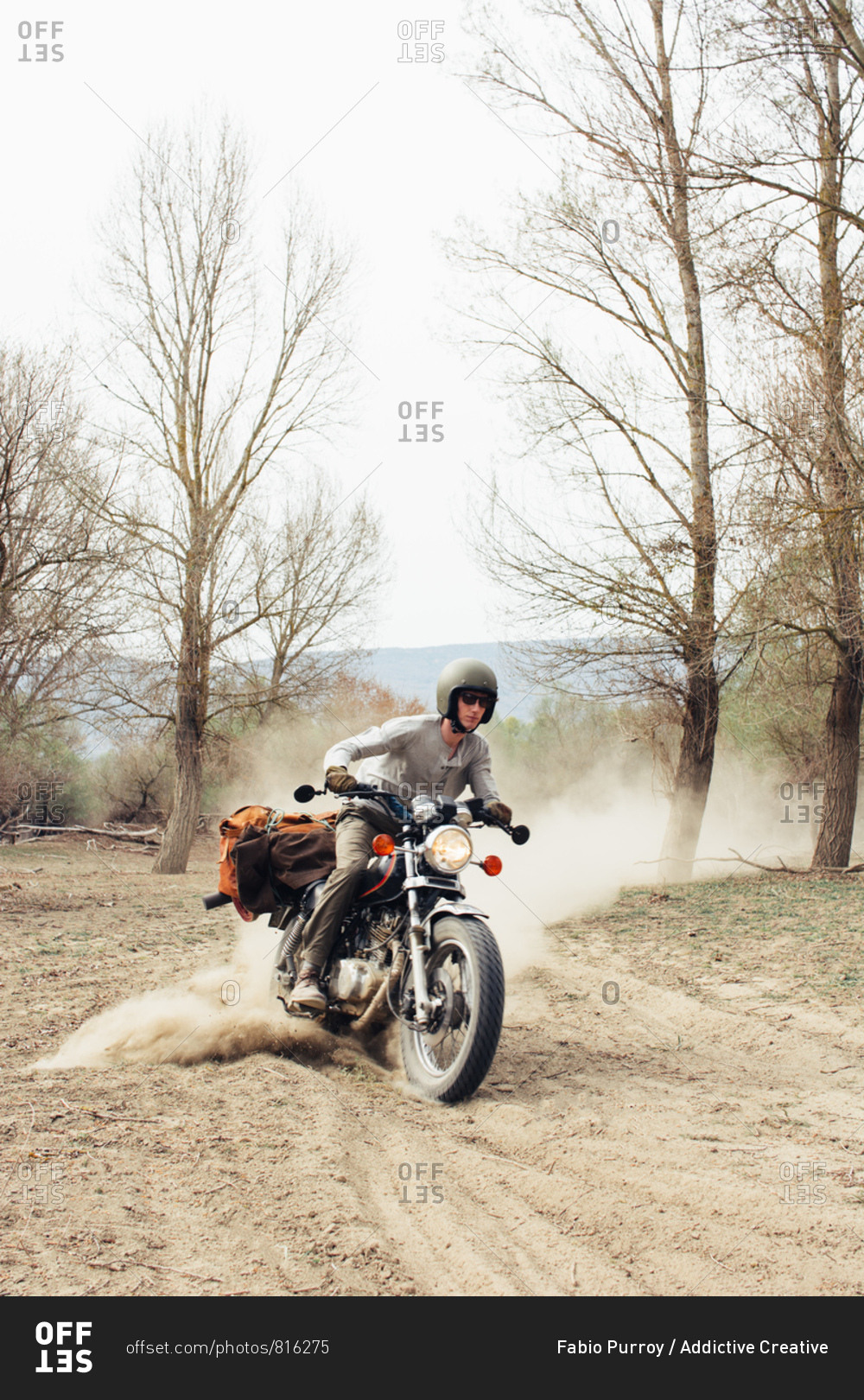 Male in helmet riding fast motorbike on dusty countryside road near leafless trees in nature