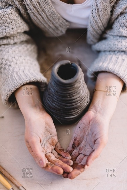 Unrecognizable potter holding dirty hands near coiled clay pot on table in workshop