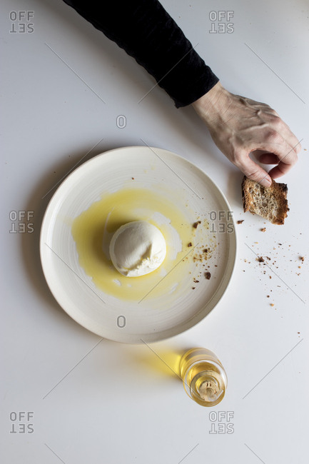 Hands of anonymous elderly person holding green napkin near plate with tasty burrata and piece of bread with oil on white background