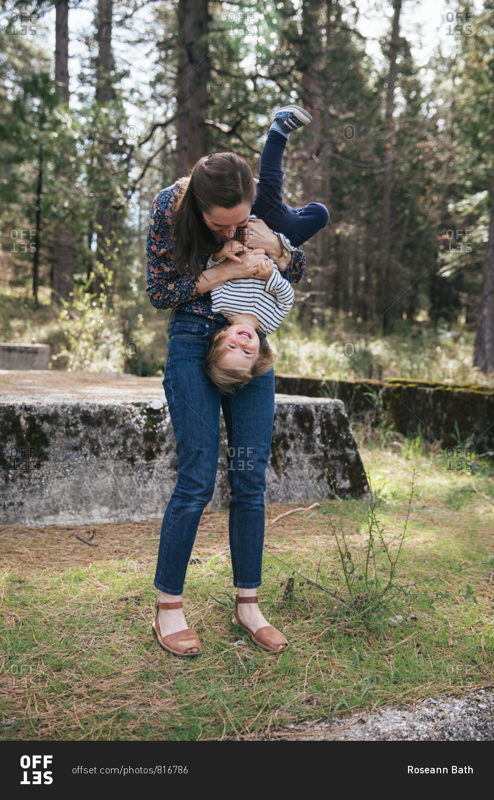 A young mother holds her toddler daughter upside down on a concrete bunker with trees in the distance
