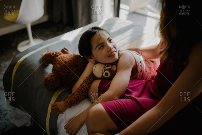 Young daughter looking up at mother while relaxing on a hotel bed with a stuffed animal monkey