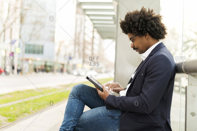Spain- Barcelona- businessman in the city sitting on bench at a station using tablet