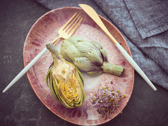 Small knife and fork and cut and whole fresh artichokes placed on pink ceramic plate on table