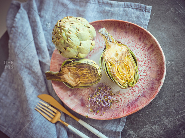 Small knife and fork and cut and whole fresh artichokes placed on pink ceramic plate on table
