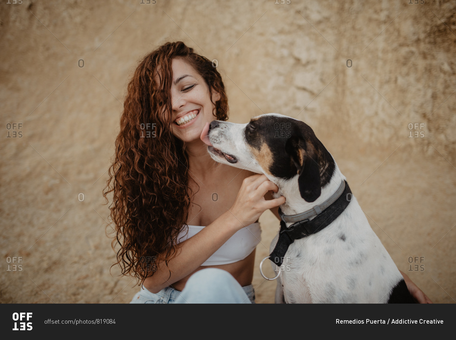 Funny dog licking cheek of excited young lady against weathered building wall on street