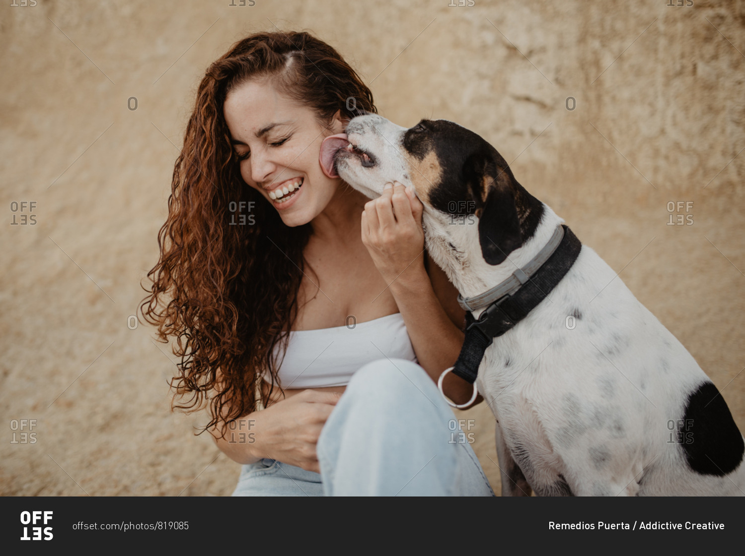 Funny dog licking cheek of excited young lady against weathered building wall on street