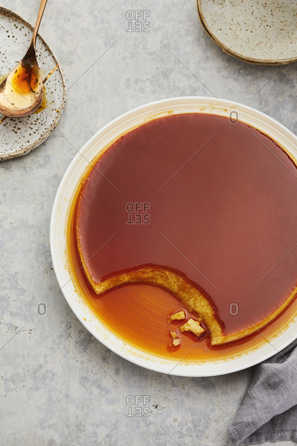 Big creme caramel flan on a plate with syrup on a marble counter top near a plate with a spoon on it.
