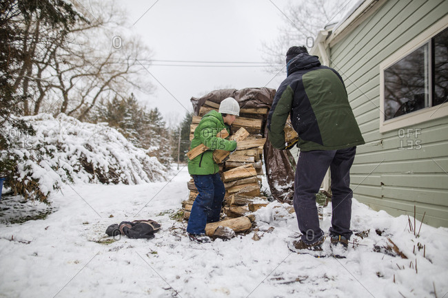 A small boy helps his father gather wood from a wood stack in the snow
