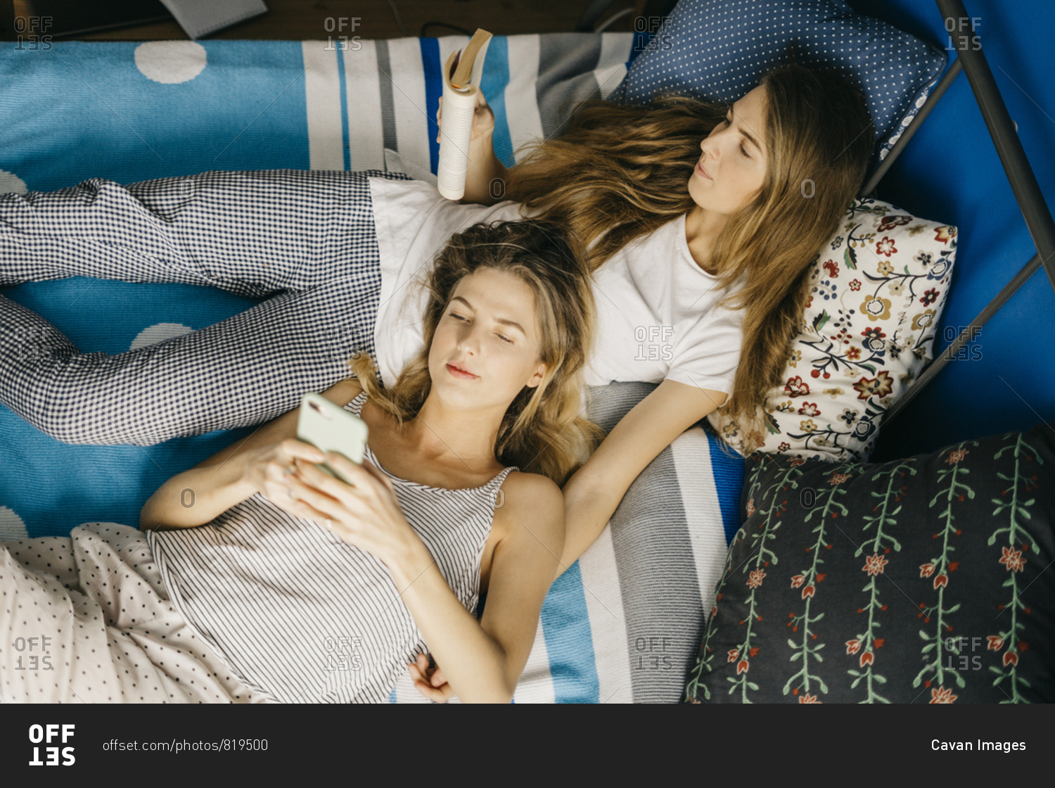 two women twins are lying on the bed, reading book, typing smartphone