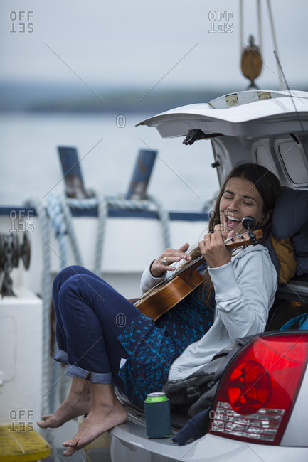 Woman singing and playing ukulele in the open trunk of her car.