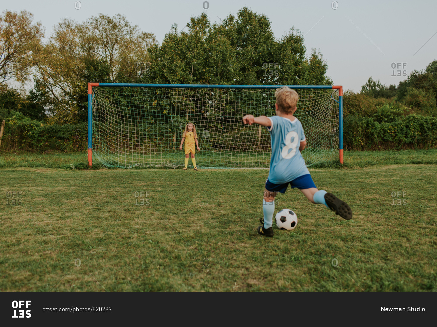 Back view of a child wearing football dress shooting ball in front of goal. Full length of girl goalkeeper standing at goal and soccer player kicking ball.