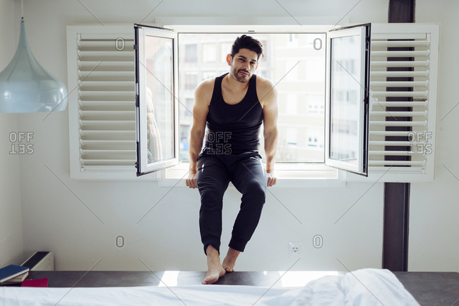Young cheerful successful male in casual singlet sitting on window near bed with books and looking at camera
