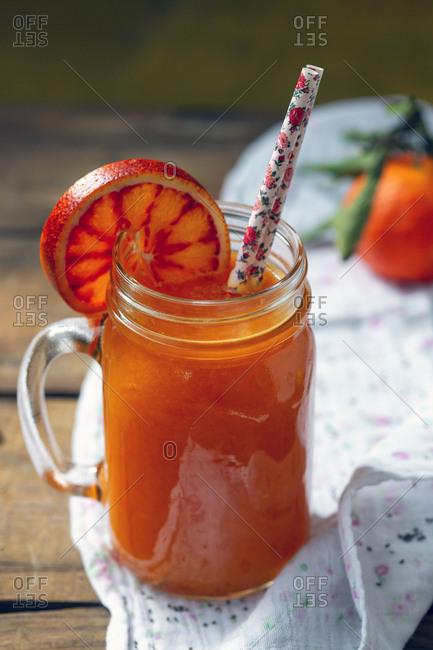 Jar of yummy fresh citrus juice with slice of ripe blood orange and straws placed on timber tabletop near napkin