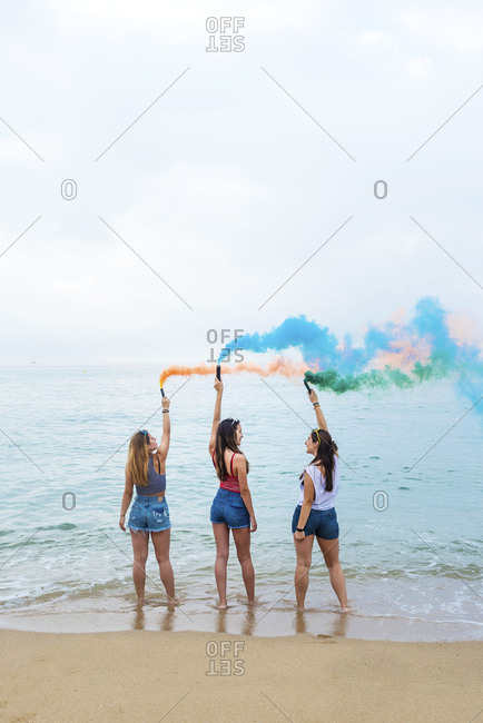 Happy teenage girls holding colored smoke bombs and having fun on the beach
