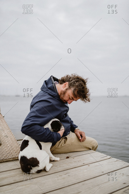 Adult male in warm jacket embracing spotted French Bulldog while sitting on wooden pier and admiring view of rippling sea on dull day