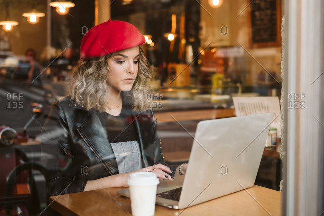 Beautiful young female in red beret drinking hot beverage window while laptop in cafe