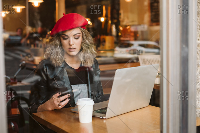 Trendy young female in red beret on mobile phone while sitting at table with laptop in restaurant