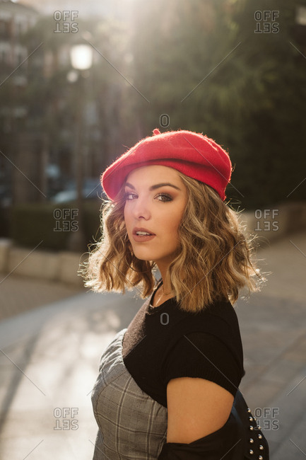 Beautiful young woman wearing trendy red beret and looking at camera while standing on city street on sunny day