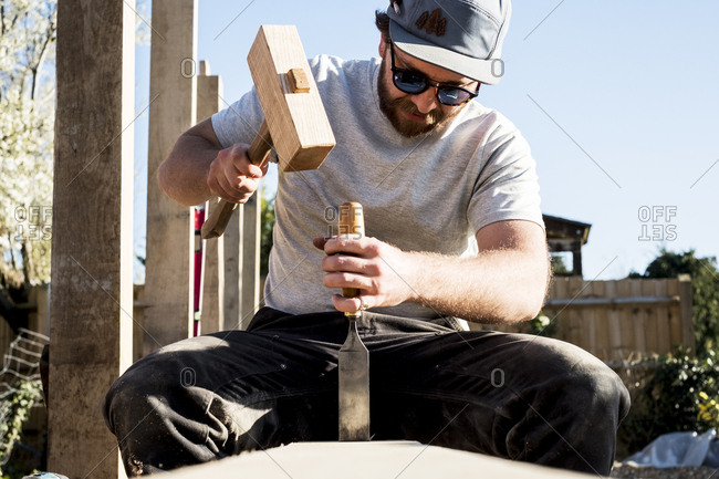 Man wearing baseball cap and sunglasses on building site, using mallet and chisel, working on wooden beam.
