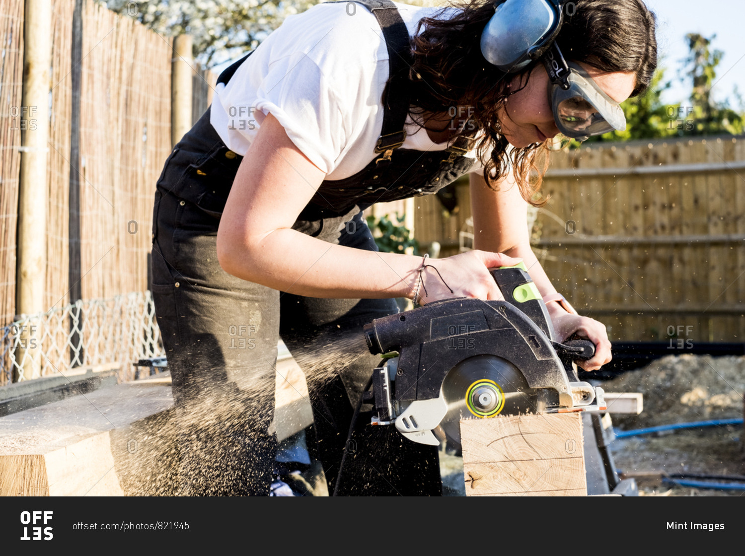 Woman wearing protective goggles and ear protectors holding circular saw, cutting piece of wood on building side.