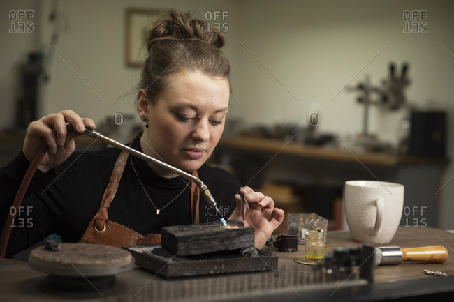 Female goldsmith heating metal with welding torch to make jewelry at workshop