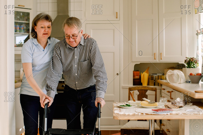 Mature female caregiver assisting retired man walking with rollator in kitchen at nursing home