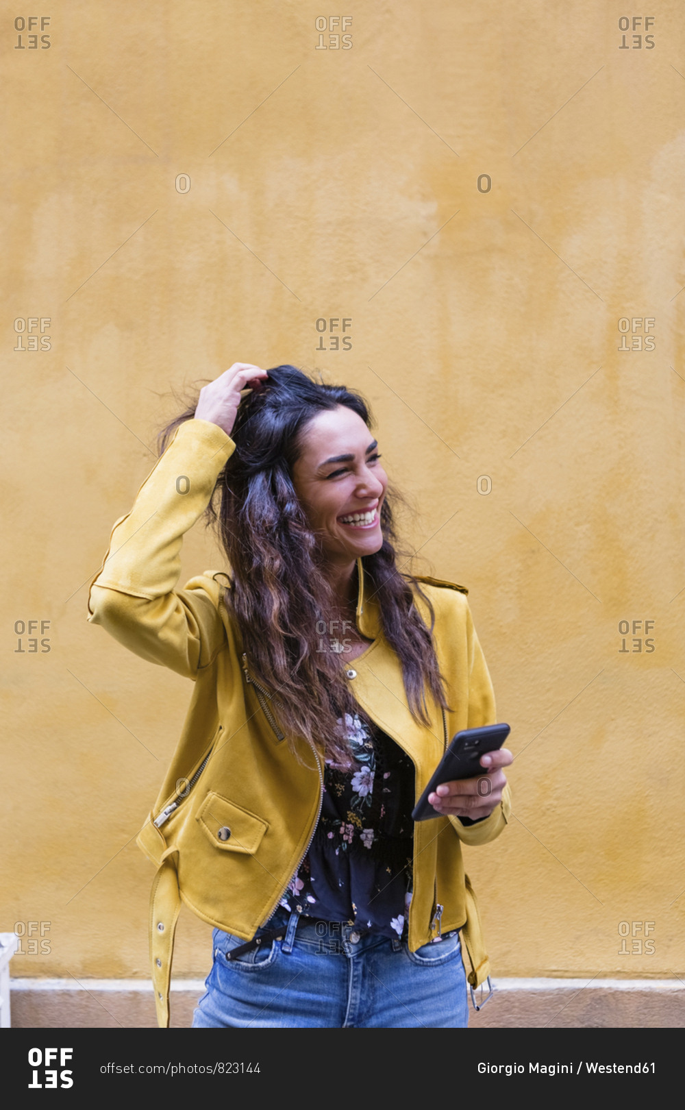 Portrait of young laughing woman wearing yellow leather jacket and holding smartphone