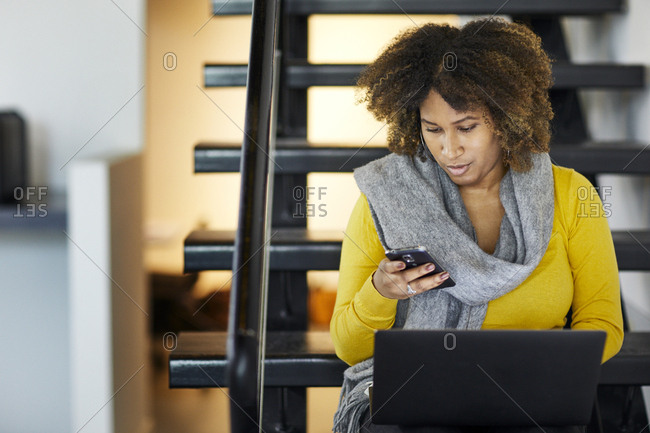 Businesswoman with laptop computer using mobile phone while sitting on steps in office