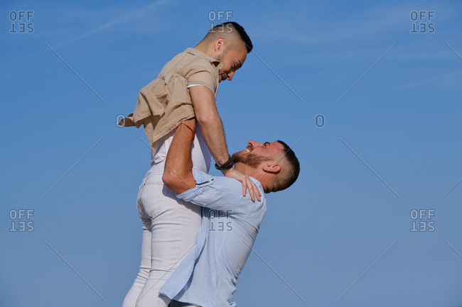 Side view of happy gay man lifting up and going to kiss boyfriend on deserted beach on summertime