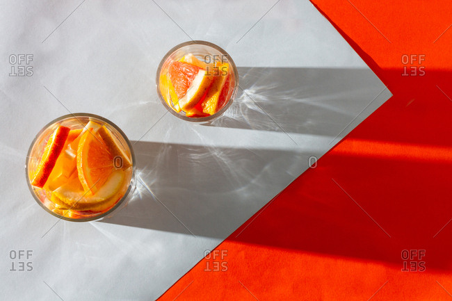 From above fresh slices orange and lemon in glass cups cast shadow on white orange background