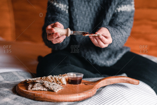 Crop view of woman hand taking delicious snack of cheese with crackers and marmalade on wooden board on bed