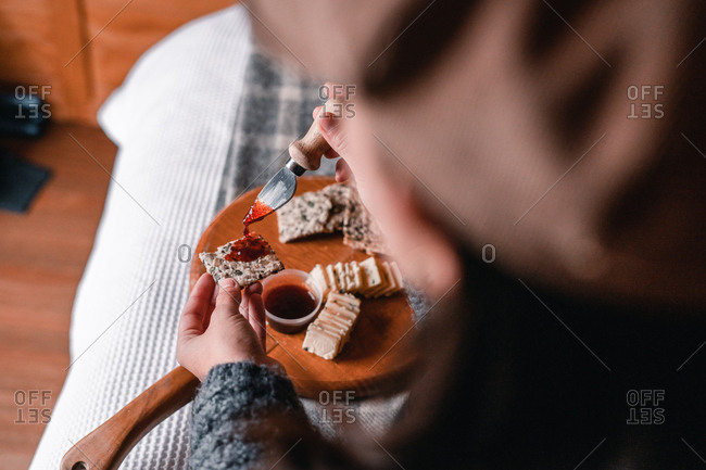 Crop view of woman hand taking delicious snack of cheese with crackers and marmalade on wooden board on bed