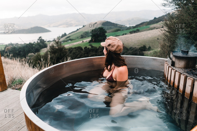 Back view of anonymous young woman in cap and bikini bathing in private wood-fired hot tub and looking at camera in Te Wepu Intrepid Pods New Zealand
