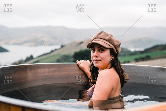 Young woman in cap and bikini bathing in private wood-fired hot tub and looking at camera in Te Wepu Intrepid Pods New Zealand