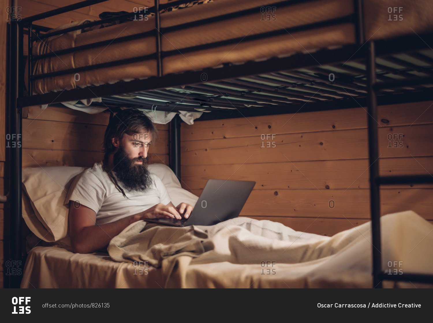 Concentrated bearded hipster working on laptop while lying in simple bunk bed of country wooden house