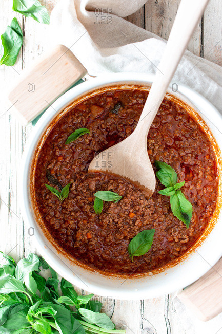 From above white ceramic pan with ragout Bolognese and spoon placed on wooden table with towel and basil