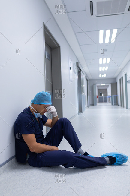 Side view of handsome tensed Caucasian male surgeon with hand over face sitting in the corridor at hospital. Surgeon is wearing surgical mask, surgical cap, gown, and surgical gloves.