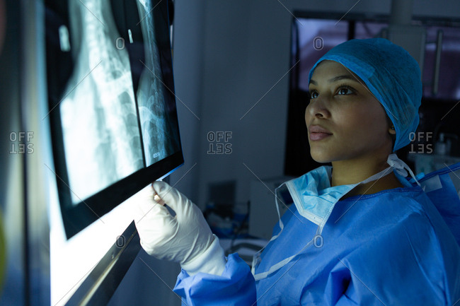 Side view of beautiful young mixed race female surgeon examining x-ray on light box in operation theater at hospital. Surgeon wears surgical gown, mask, cap and gloves.