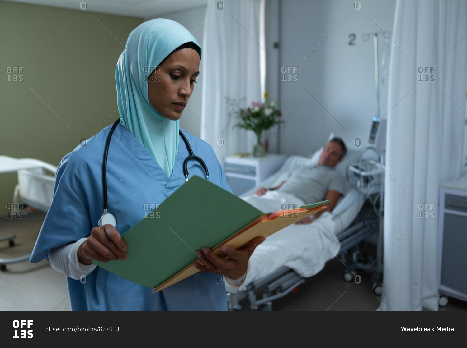 Front view of mixed race female doctor with hijab looking at medical report while Caucasian male patient sleeps in bed in the ward in hospital