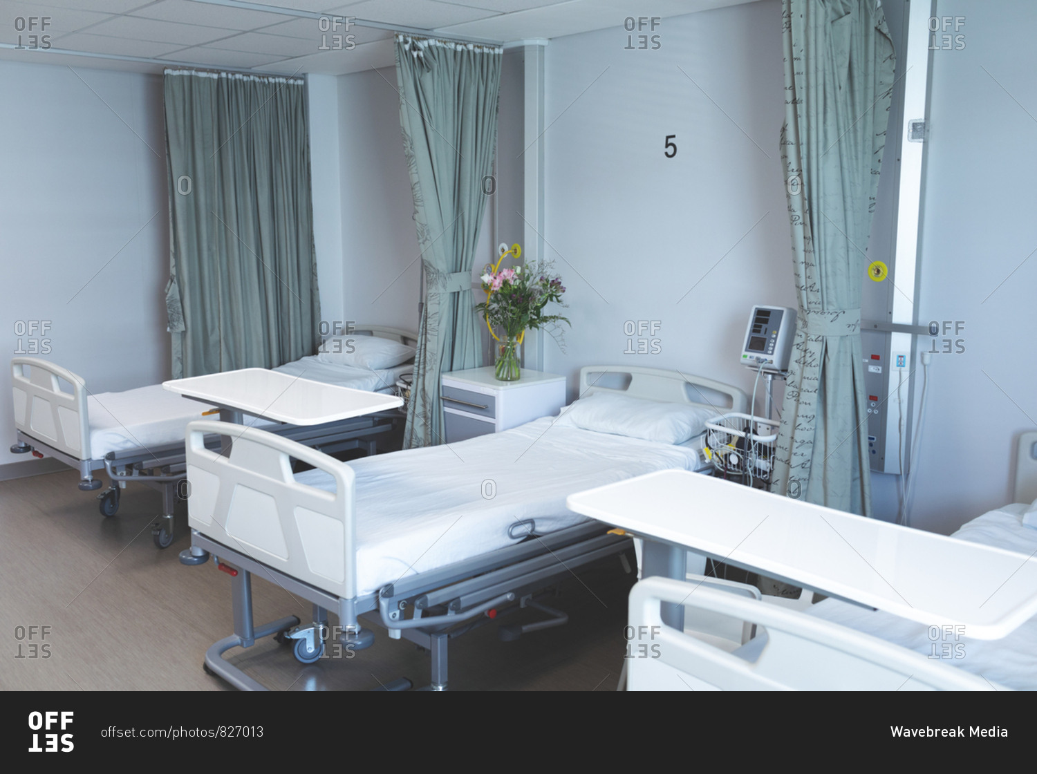 Modern hospital ward with empty beds, medical monitor, green curtains, cupboards, and flowers.