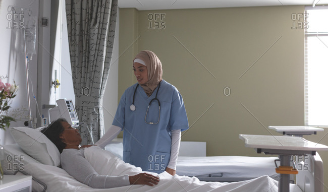Side view of beautiful mixed-race female nurse in hijab interacting with mixed-race female patient in the ward at hospital. Nurse has stethoscope around her neck.