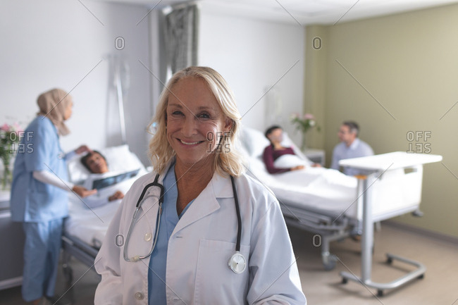 Portrait of Caucasian female doctor smiling in the ward at hospital. In the background diverse doctors are interacting with their patients next to the beds.