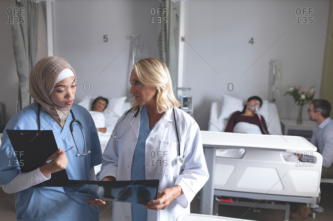 Front view of diverse female doctors discussing over x-ray report in the ward at hospital. In the background diverse doctors are interacting with their patients.
