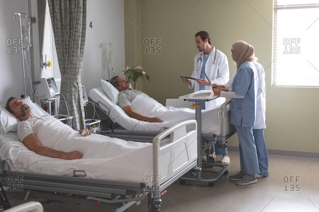 Side view of diverse doctors interacting with senior mixed-race male patient while Caucasian male patient sleeping in bed next to them in the ward at hospital