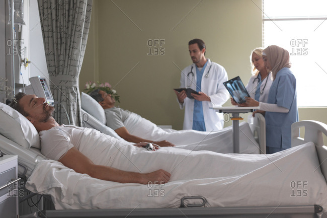 Side view of diverse doctors interacting with mixed-race male patient  while Caucasian male patient sleeping in bed next to them  in the ward at hospital.