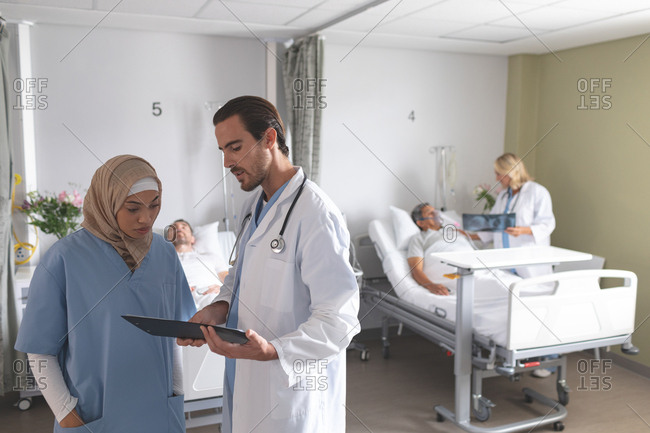 Front view of diverse doctors discussing over clipboard in the ward at hospital. In the background Caucasian female doctor talks with senior mixed-race male patient about x-ray.