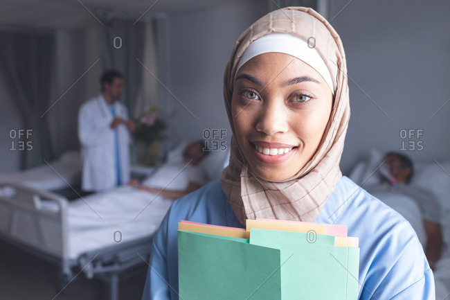 Portrait of mixed-race female doctor in hijab standing with medical files in the ward at hospital. In the background Caucasian doctor talks with patient.