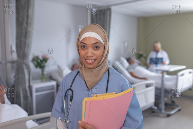 Portrait of mixed race female doctor in hijab standing with medical files in the ward at hospital. In the background diverse patients lying in bed and Caucasian female doctor checks on them.