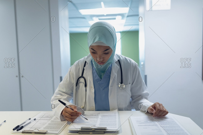Front view of mixed race female doctor in hijab reading documents at reception in hospital